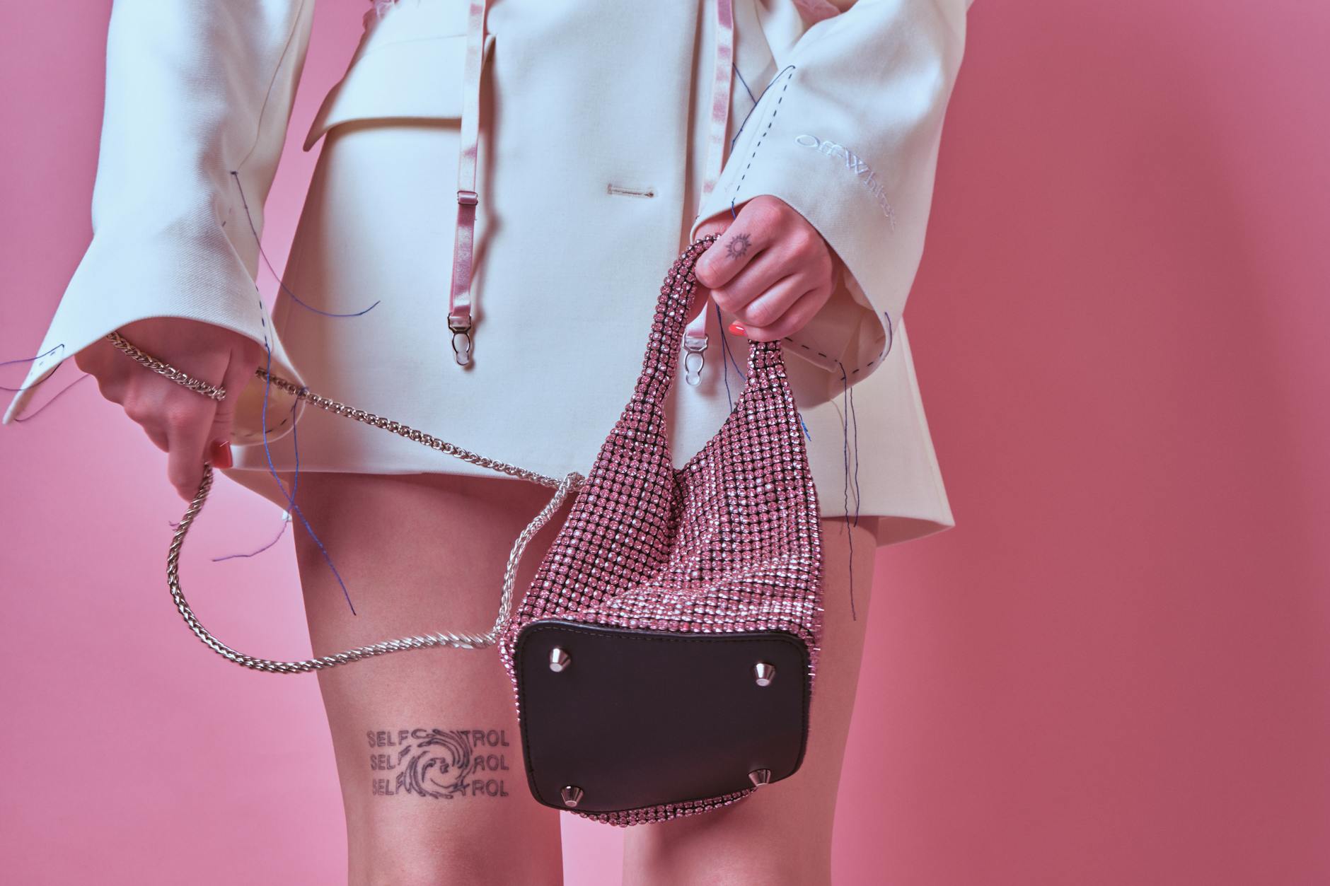 Fashion-forward image of a woman in white attire holding a pink bejeweled handbag against a pink background.
