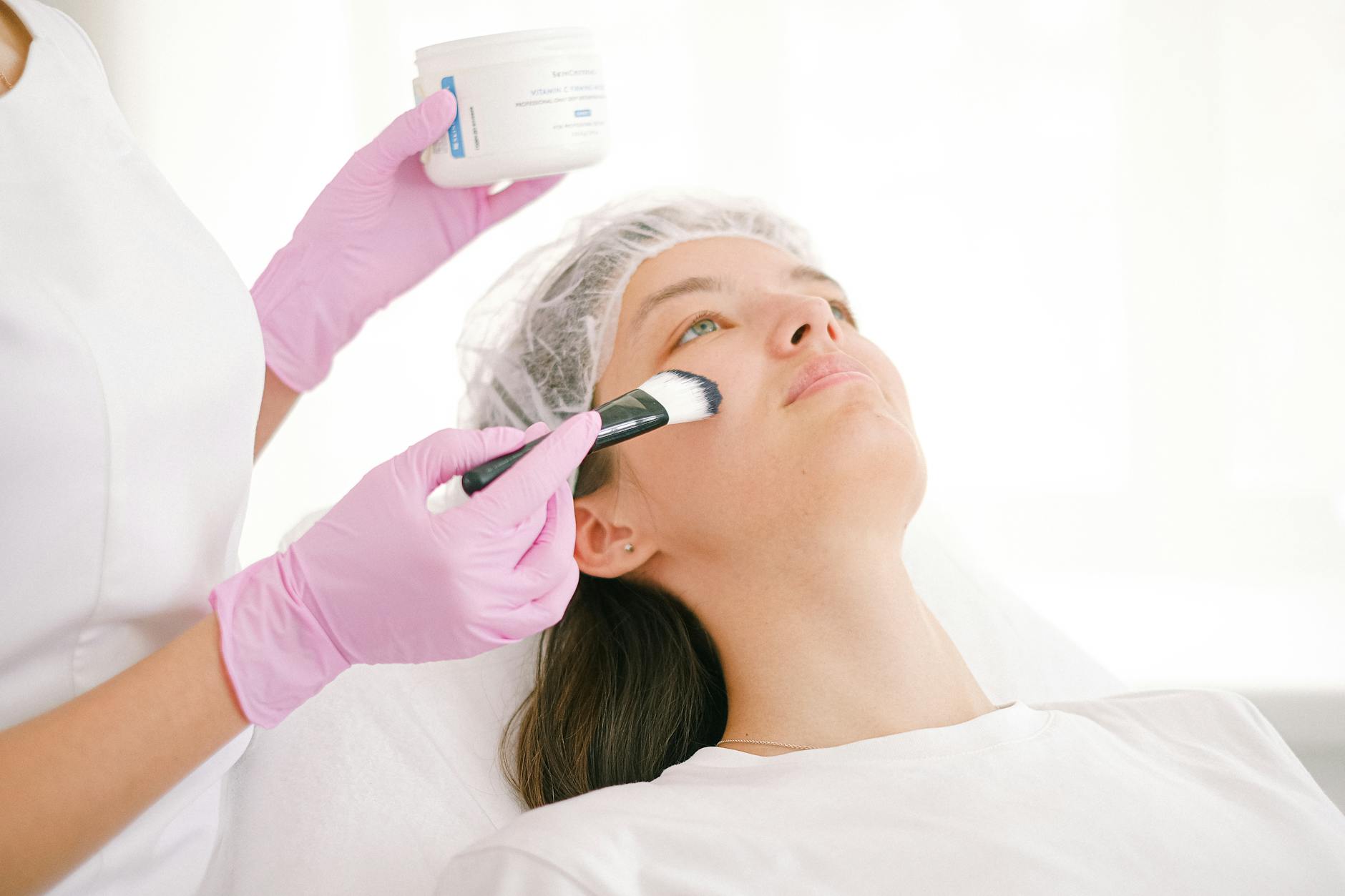A woman undergoing a facial treatment in a clinic by a professional using a brush and ointment.