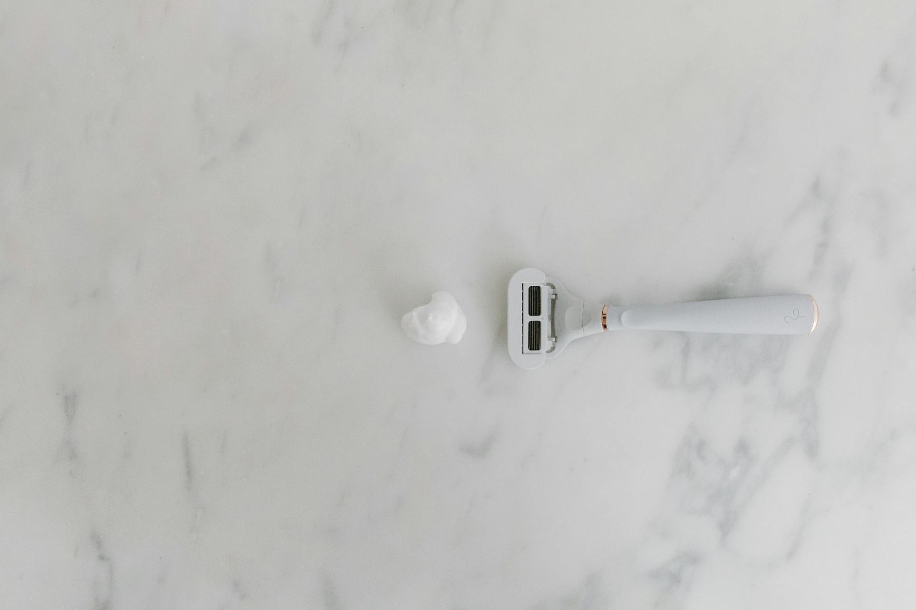 A minimalist scene with a shaving razor and foam on a marble background, offering copyspace.