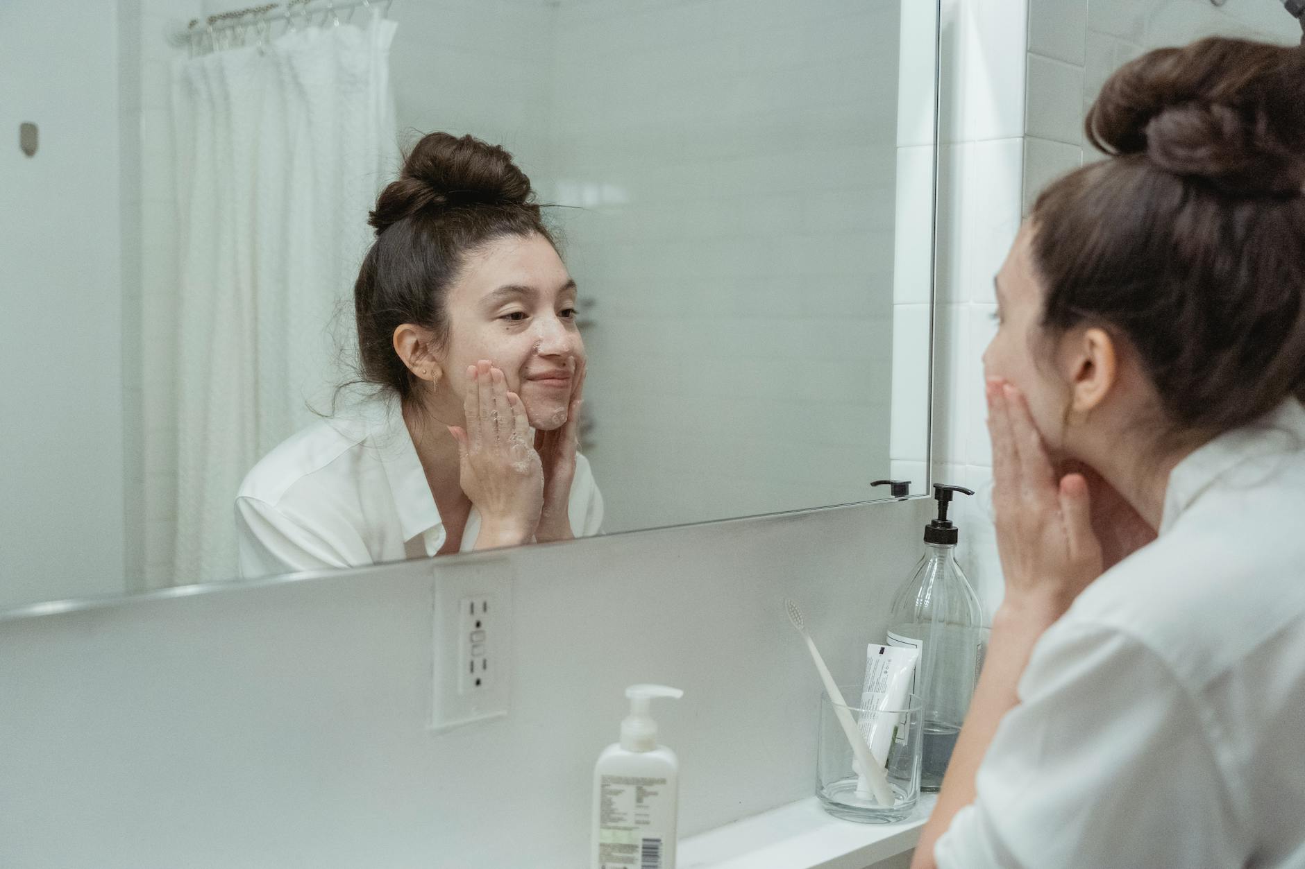Woman performing skincare routine in a bathroom, applying face cream while looking into a mirror.