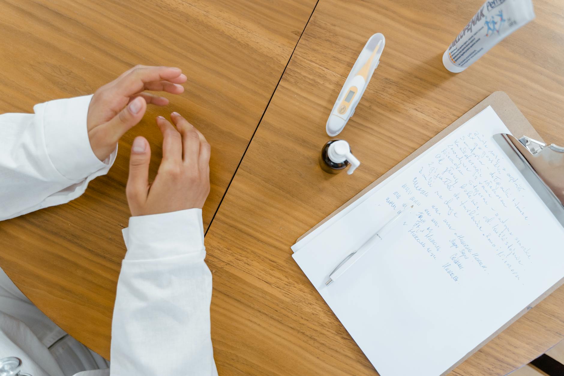 Top-down view of a doctor's desk featuring notes, thermometer, and hands in a healthcare setting.